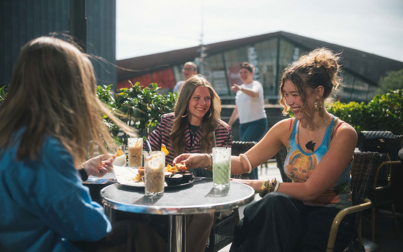 Tre unge kvinder sidder udenfor en café og snakker. Der er med og drikke på bordene imellem dem.