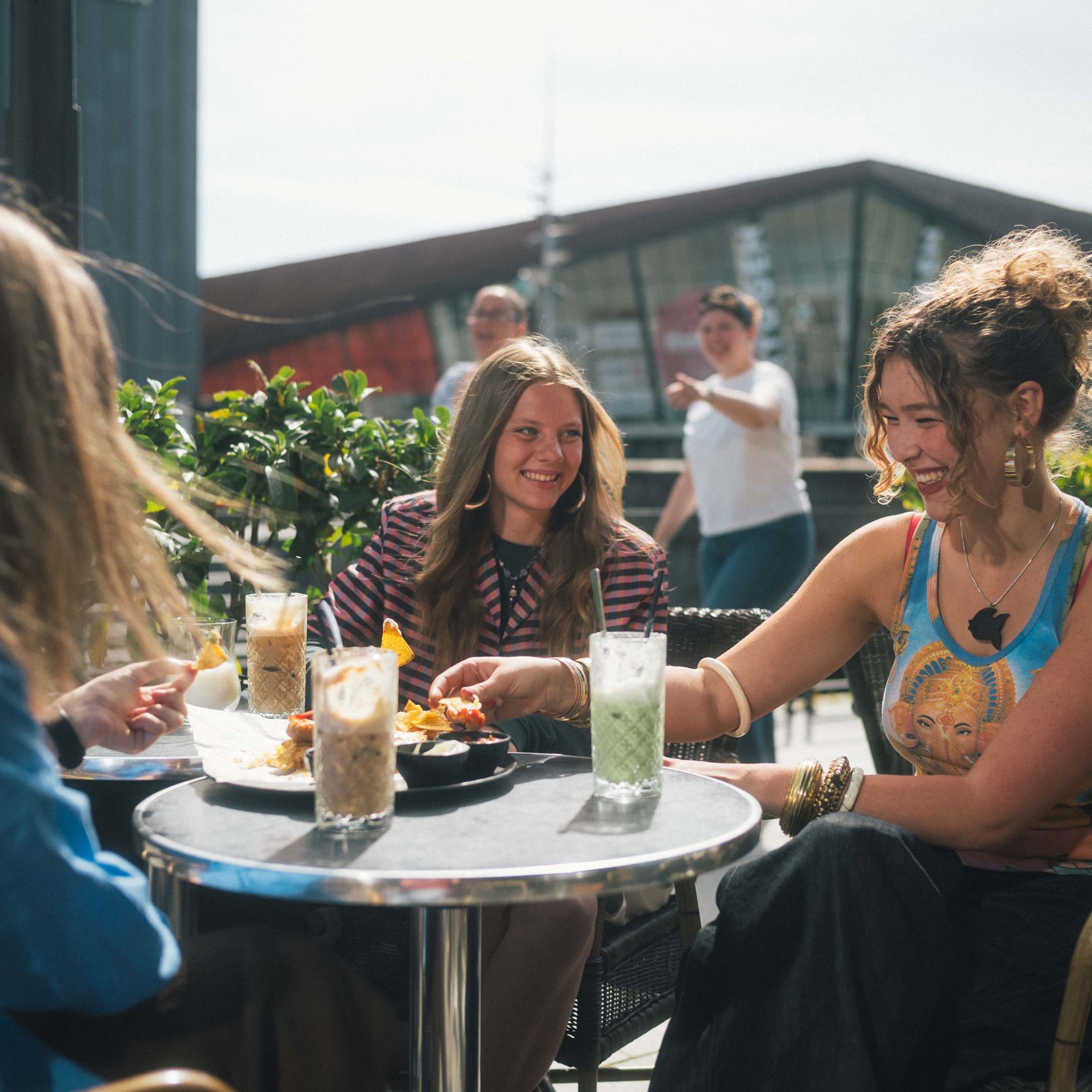 Tre unge kvinder sidder udenfor en café og snakker. Der er med og drikke på bordene imellem dem.