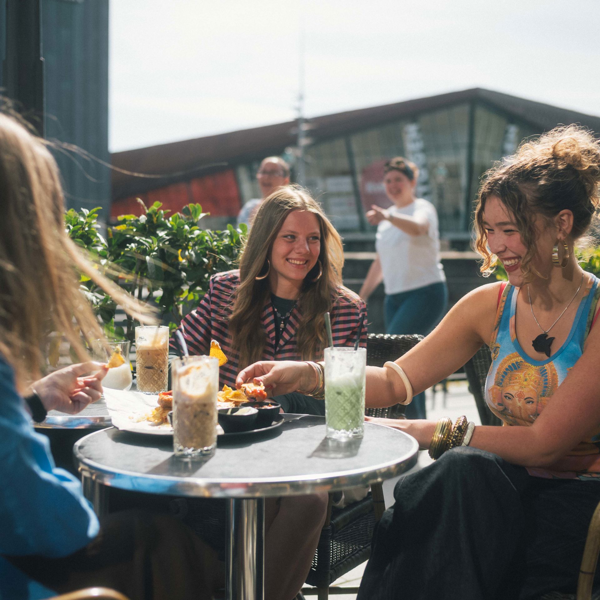 Tre unge kvinder sidder udenfor en café og snakker. Der er med og drikke på bordene imellem dem.