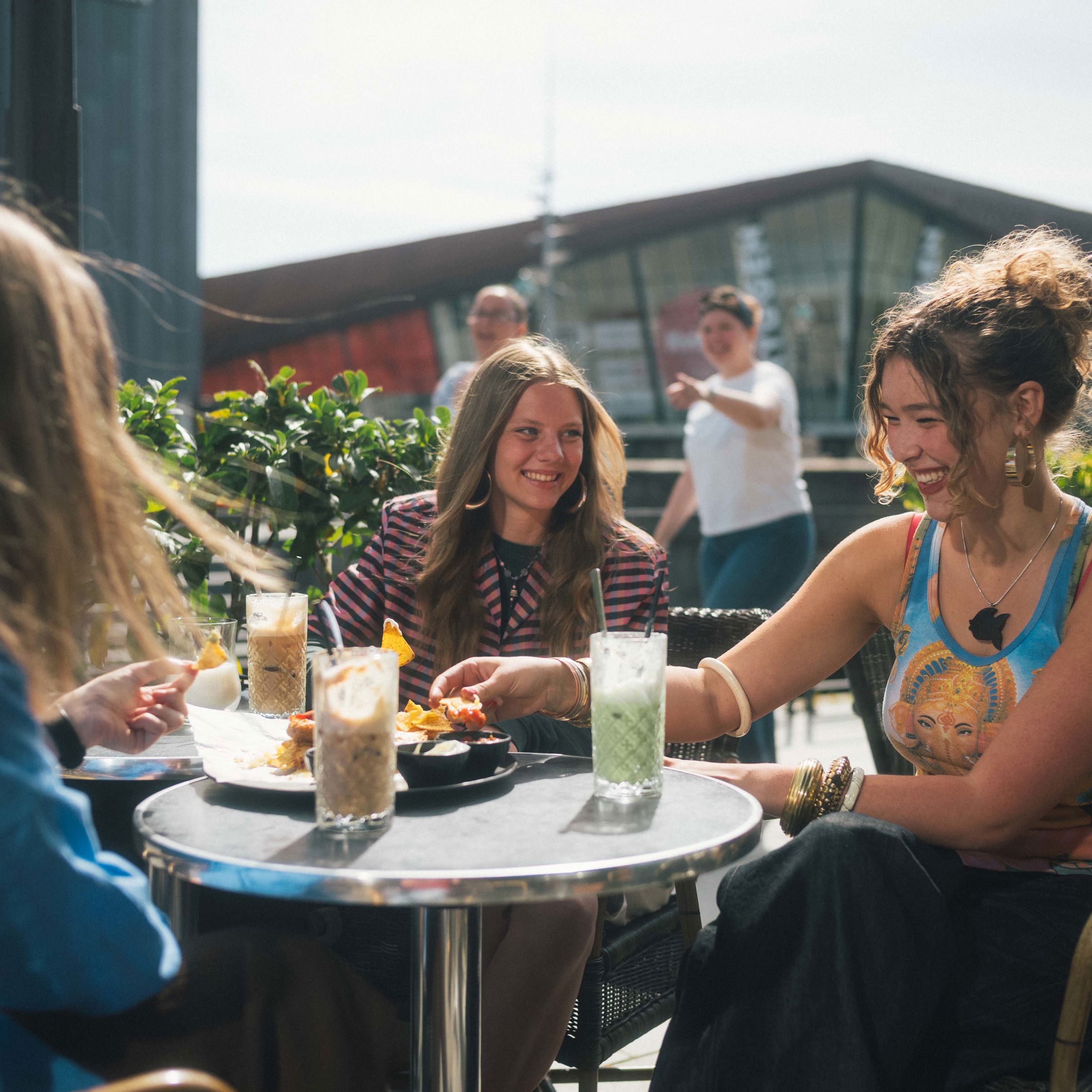 Tre unge kvinder sidder udenfor en café og snakker. Der er med og drikke på bordene imellem dem.