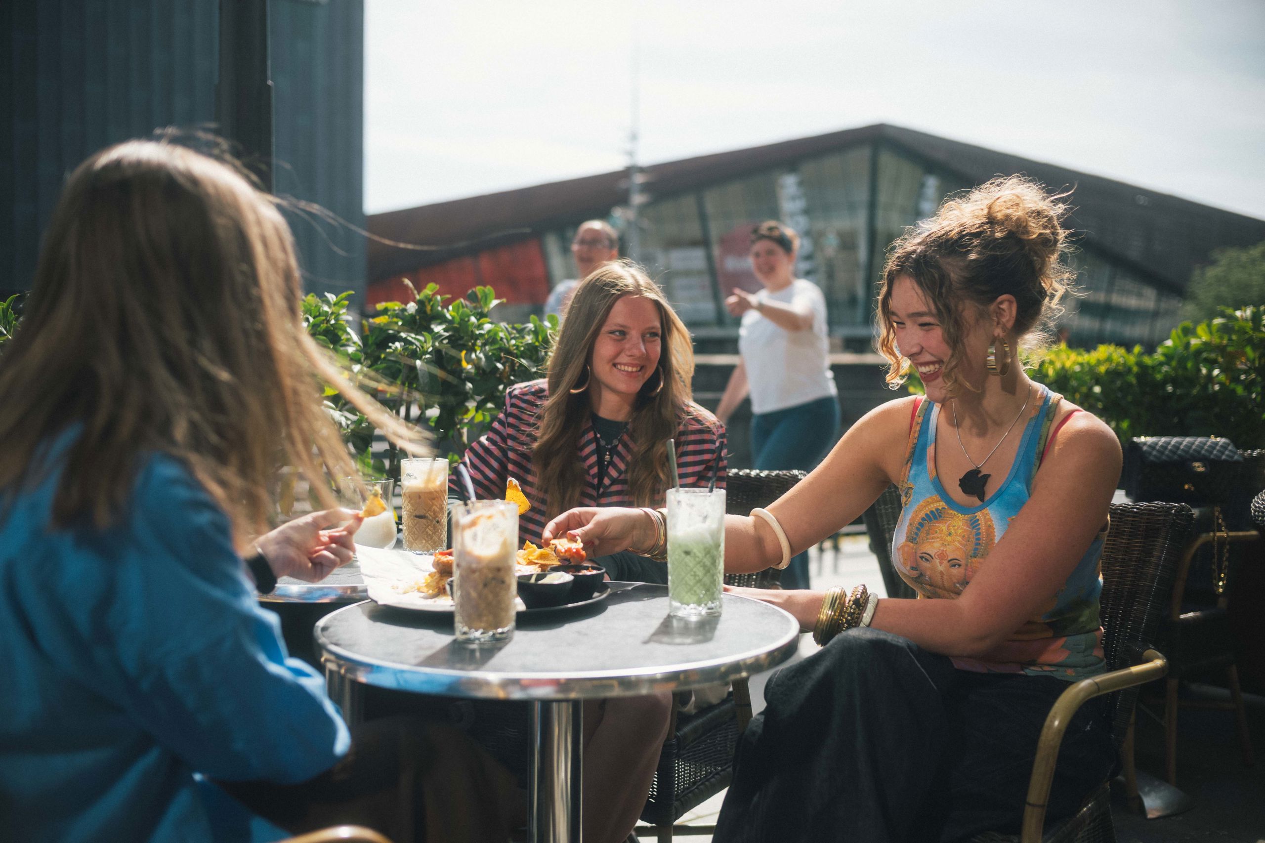 Tre unge kvinder sidder udenfor en café og snakker. Der er med og drikke på bordene imellem dem.