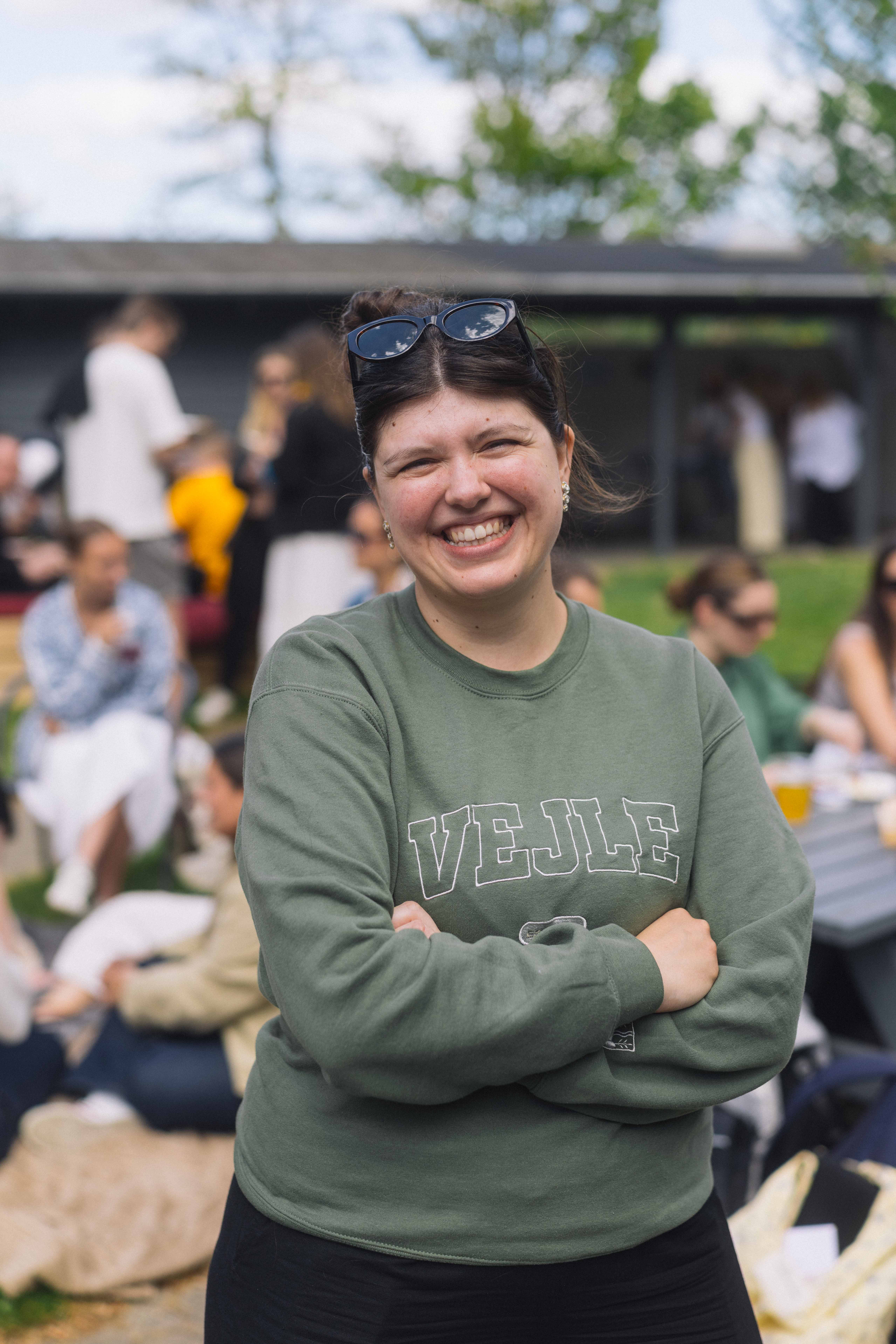 Ung kvinde smiler til kameraet. Hun har armene over kors solbriller i håret. På hendes sweatshirt står der "VEJLE".