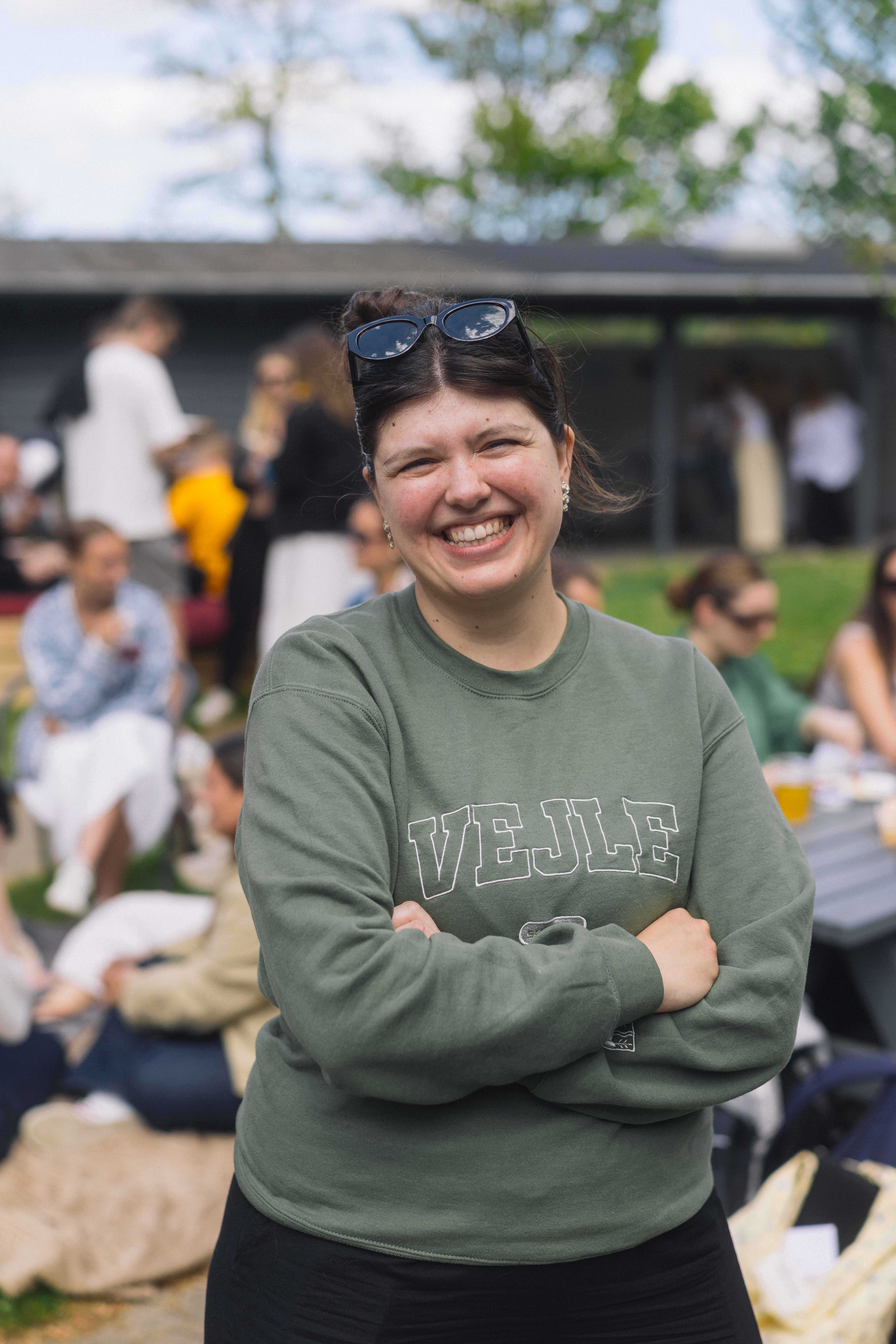 Ung kvinde smiler til kameraet. Hun har armene over kors solbriller i håret. På hendes sweatshirt står der "VEJLE".