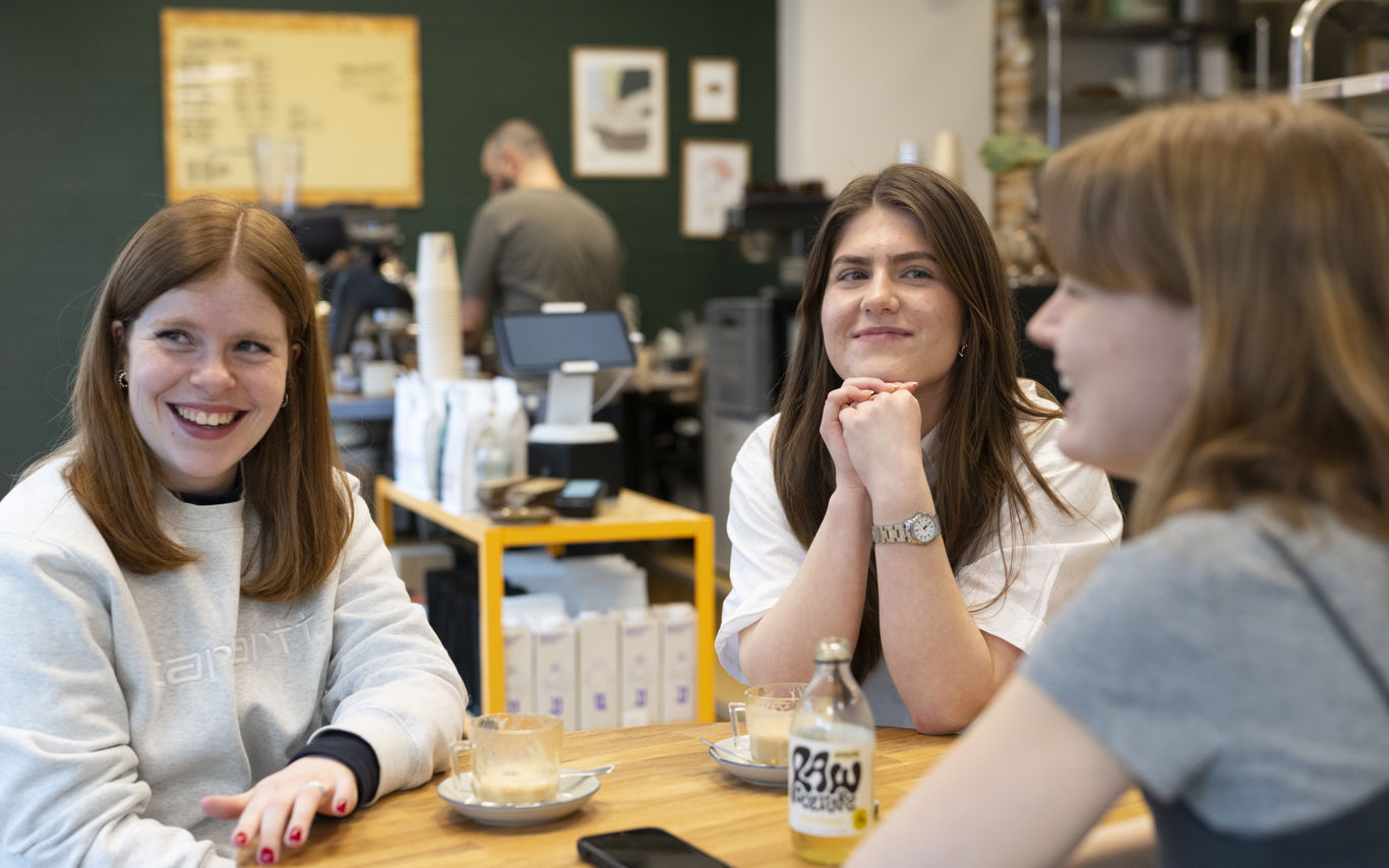 Tre unge kvinder sidder omkring et cafébord og snakker sammen.