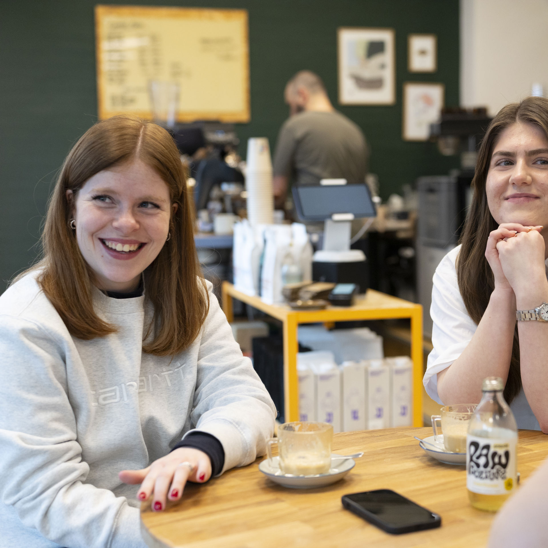 Tre unge kvinder sidder omkring et cafébord og snakker sammen.
