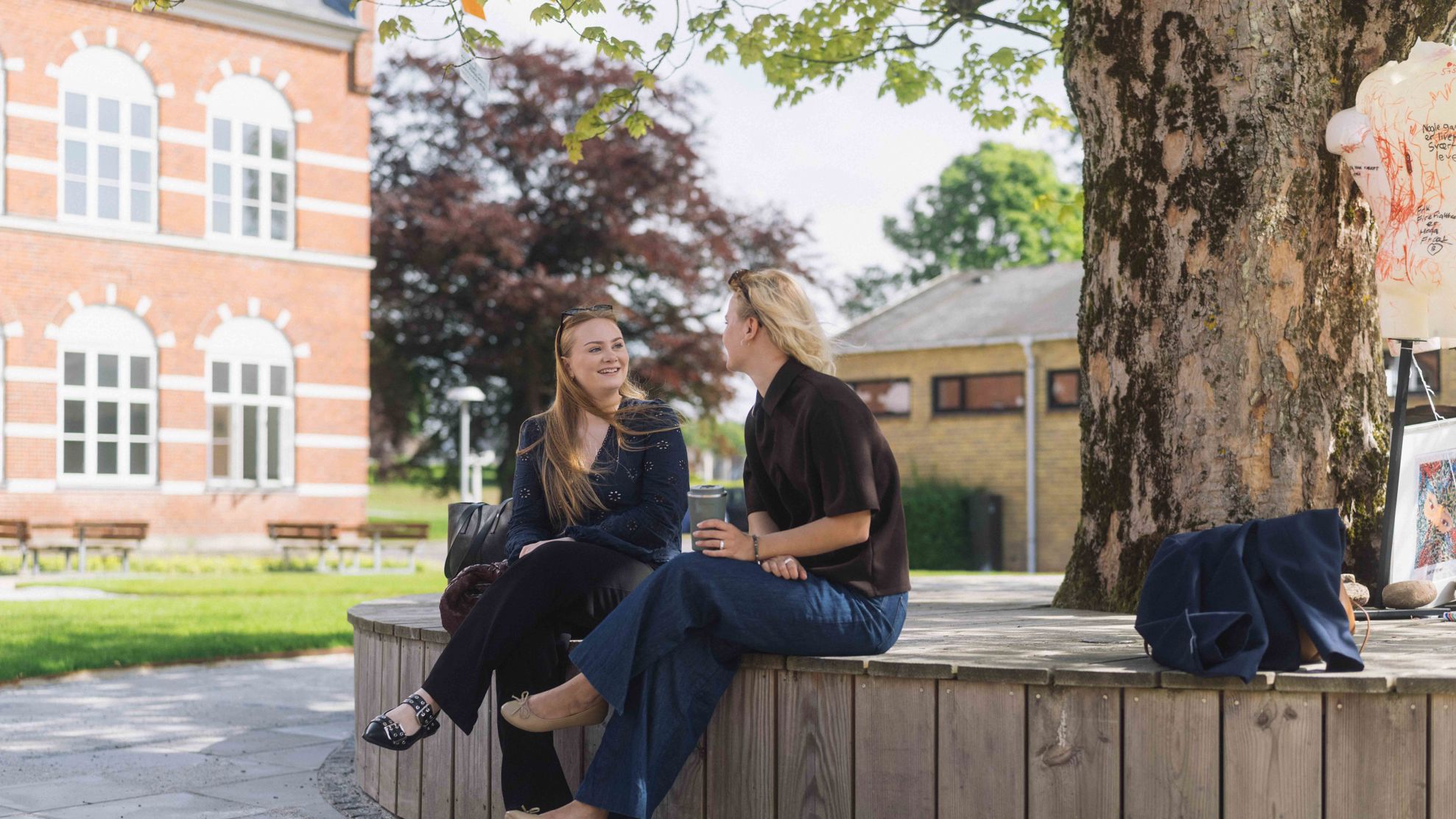 To unge kvinder sidder og snakker foran bygningerne til UCL Jelling.